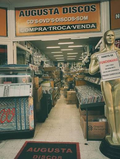 The entrance to Augusta Discos under its orange sign, a narrow aisle lined floor to ceiling with CDs and records, a golden statue by the door.