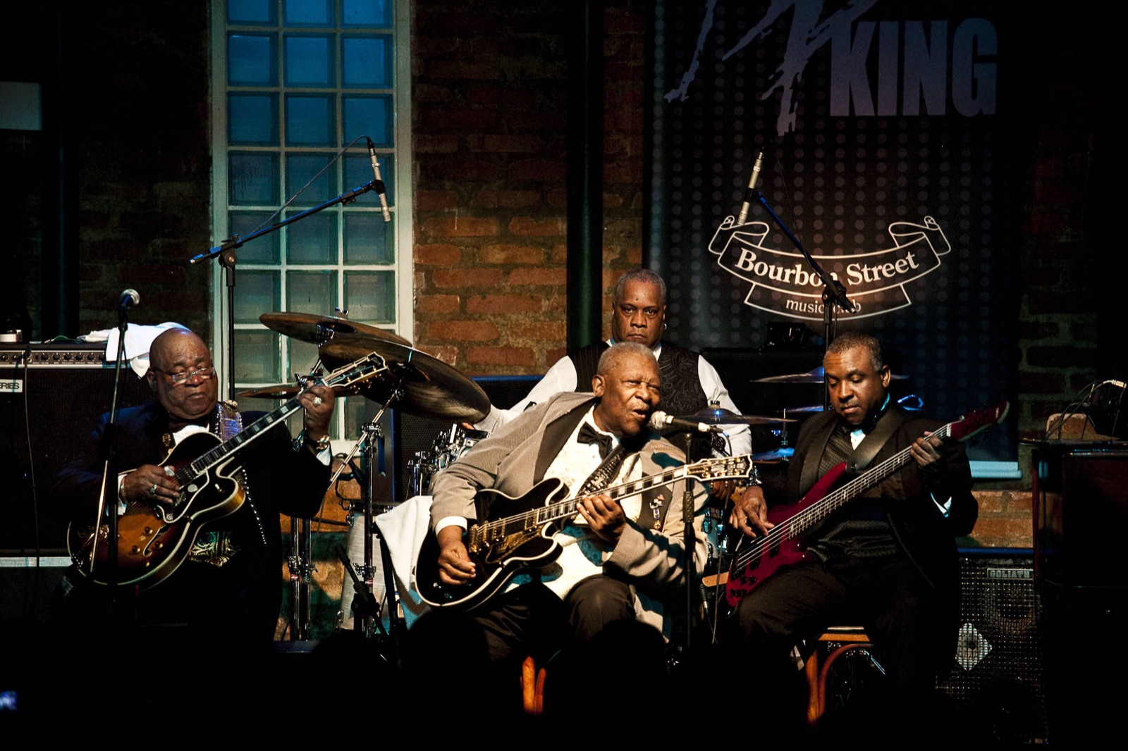 A blues band playing on the Bourbon Street Music Club stage, guitars and bass in warm light against a brick wall and the club’s banner.
