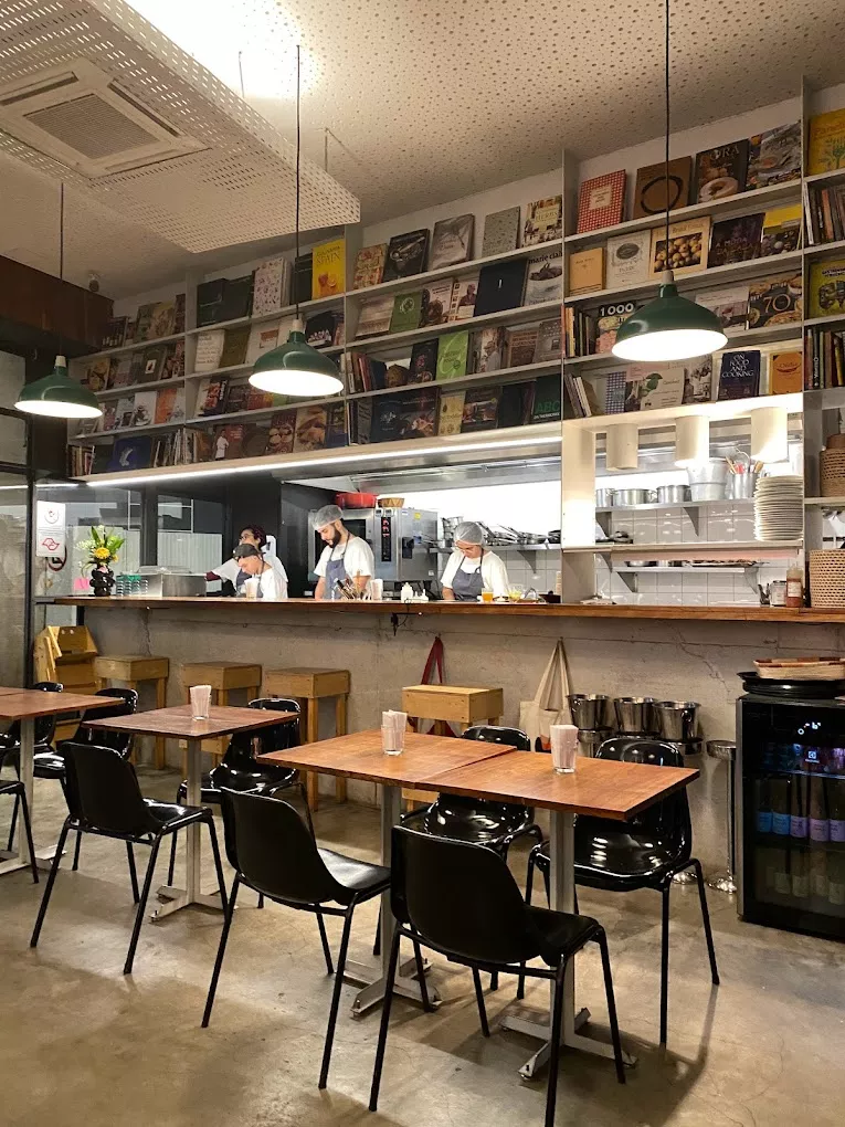 Cuia’s open kitchen behind a long wooden counter, cooks at work beneath shelves of cookbooks and green pendant lamps, empty tables in the foreground.