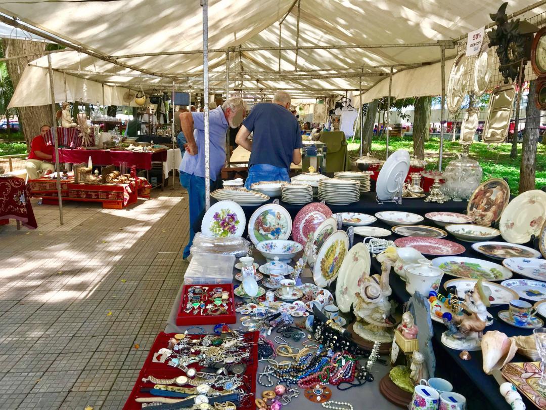A stall at the Bixiga fair under a white canopy: painted porcelain plates, teacups, figurines, and trays of costume jewelry and old watches, with people browsing behind.