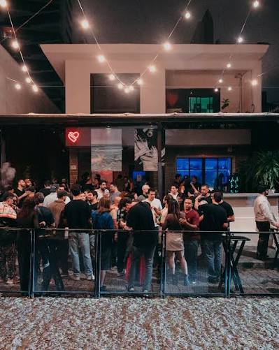 A crowd gathered outside Heavy House at night under strings of warm bulbs, a red neon heart glowing in the window behind them.
