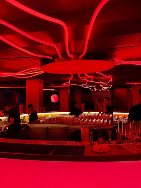 The bar at Iccarus bathed in red light, neon lines curling across the ceiling around a round light like a sun, bartenders working behind rows of glasses.