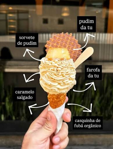 A hand holding Tem Umami’s pudim ice cream in an organic cornmeal cone, topped with a piece of pudim, a waffle crisp, crumble and salted caramel, each part labelled in Portuguese.