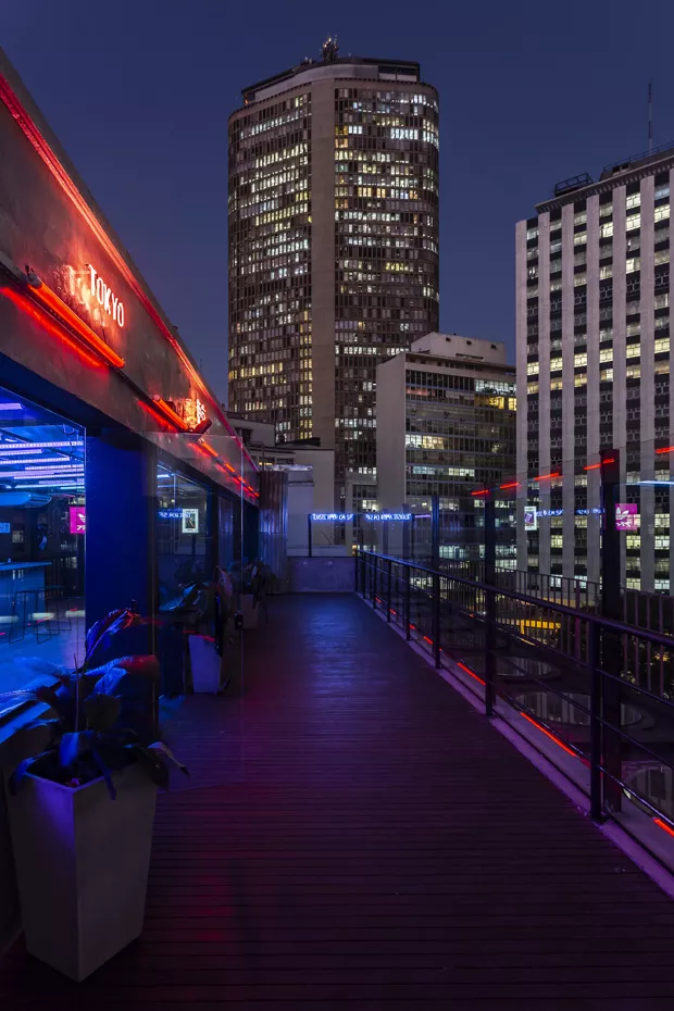 Tokyo’s rooftop deck at night, red neon along the bar and lit office towers of the city centre beyond the railing.