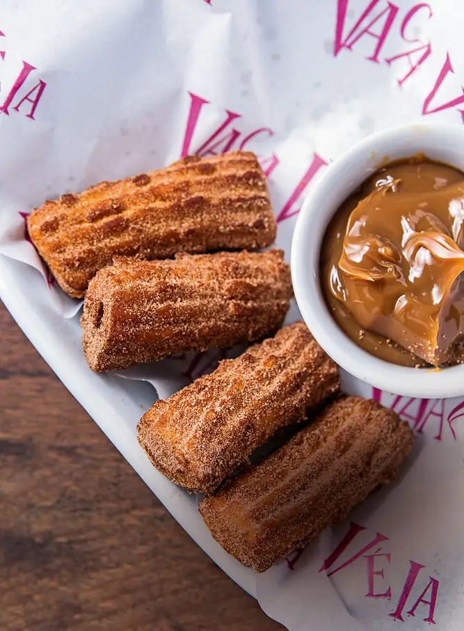 Cinnamon-sugar churros on Vaca Véia’s printed paper, with a small bowl of doce de leite for dipping.